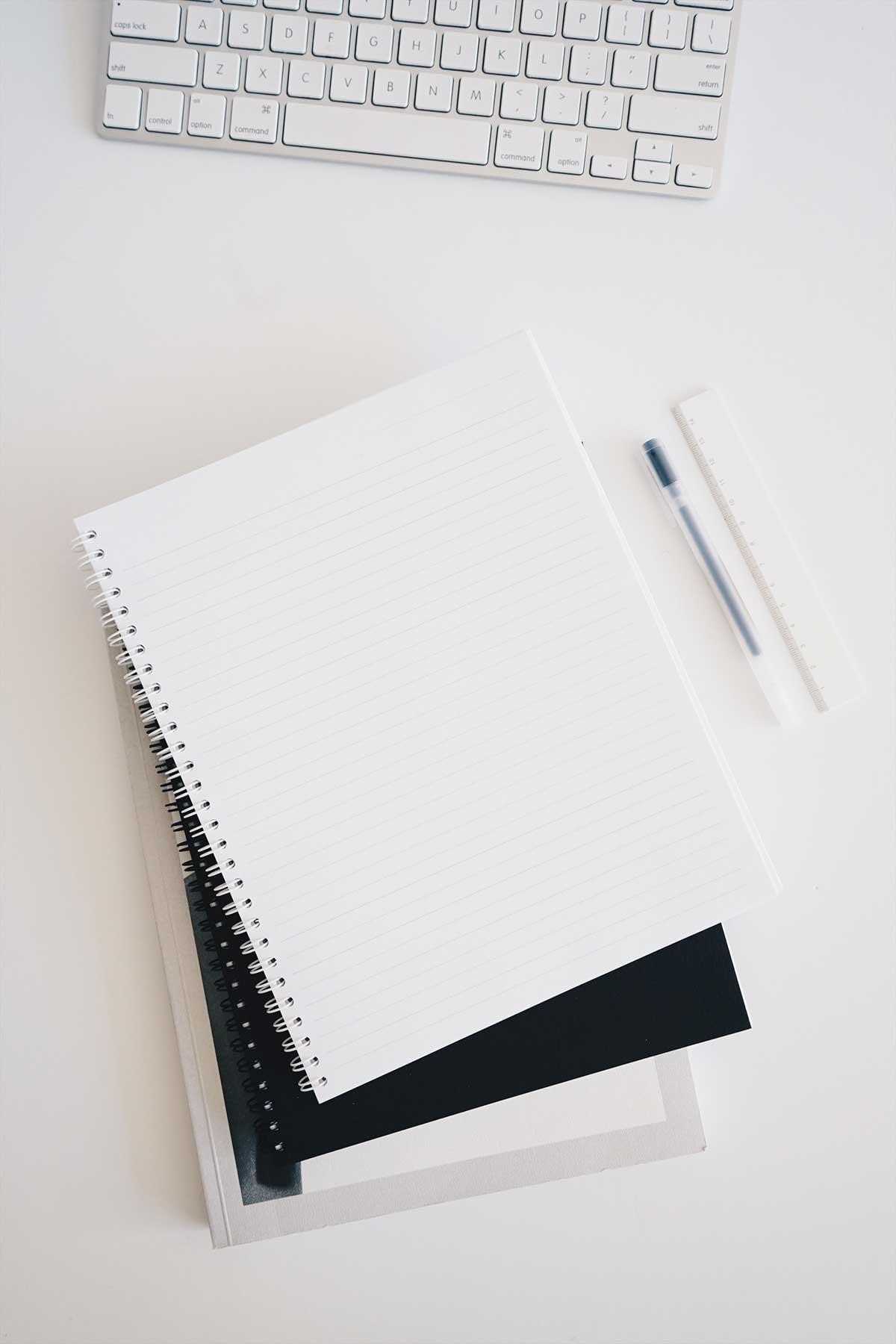 A Mac keyboard and notebook on a white table, with a pen and ruler